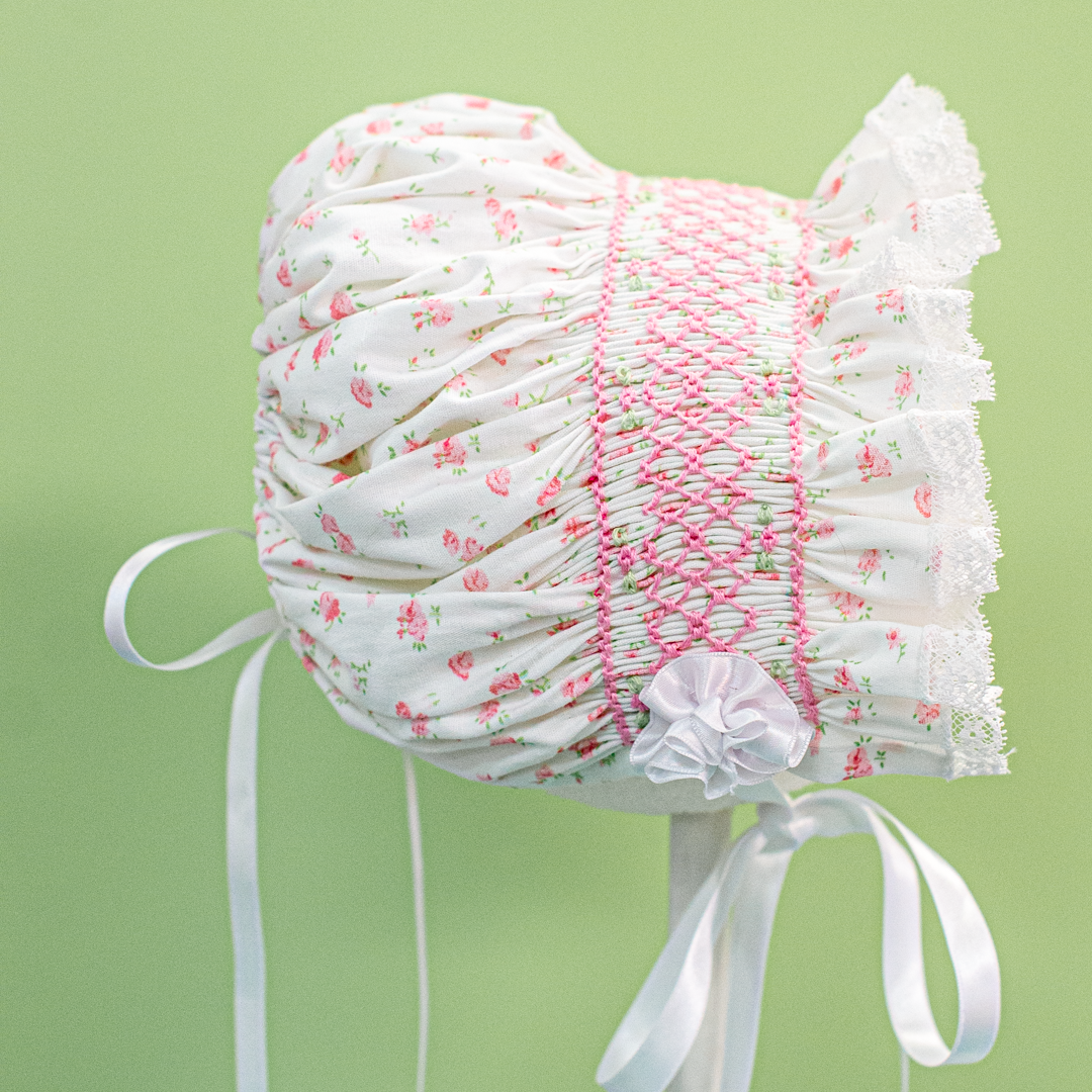 Baby bonnet in cream fabric with pink floral print, smocking, lace trim, and ribbon ties.