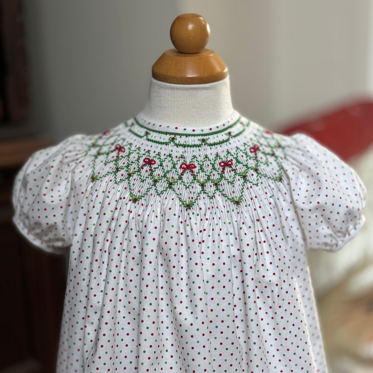 White dress with red polka dots and green smocking on a mannequin against a neutral background