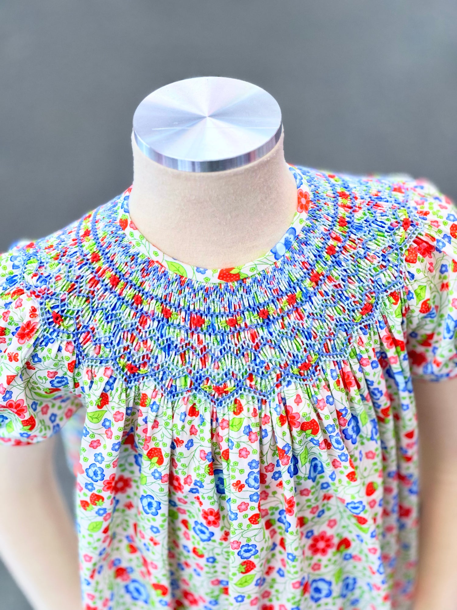 Children's smocked bishop dress in floral strawberry print with blue smocking on mannequin