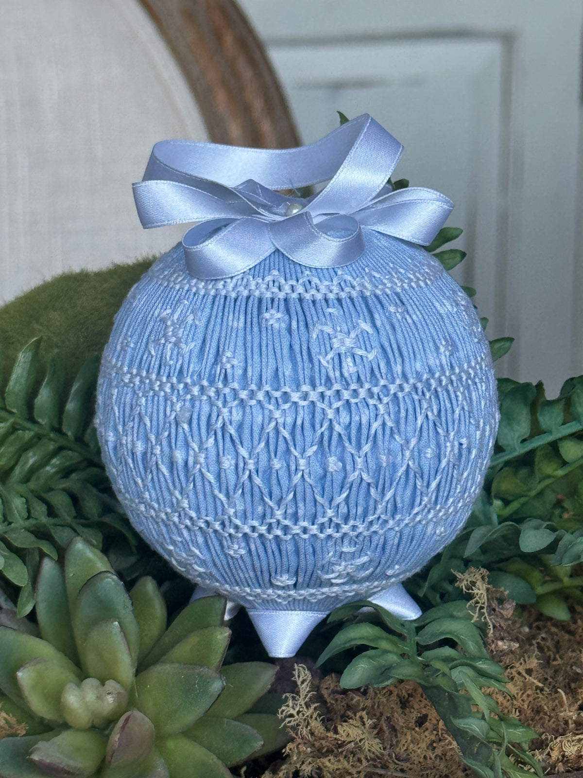Light blue smocked ornament with white embroidery, satin bow, displayed among succulents