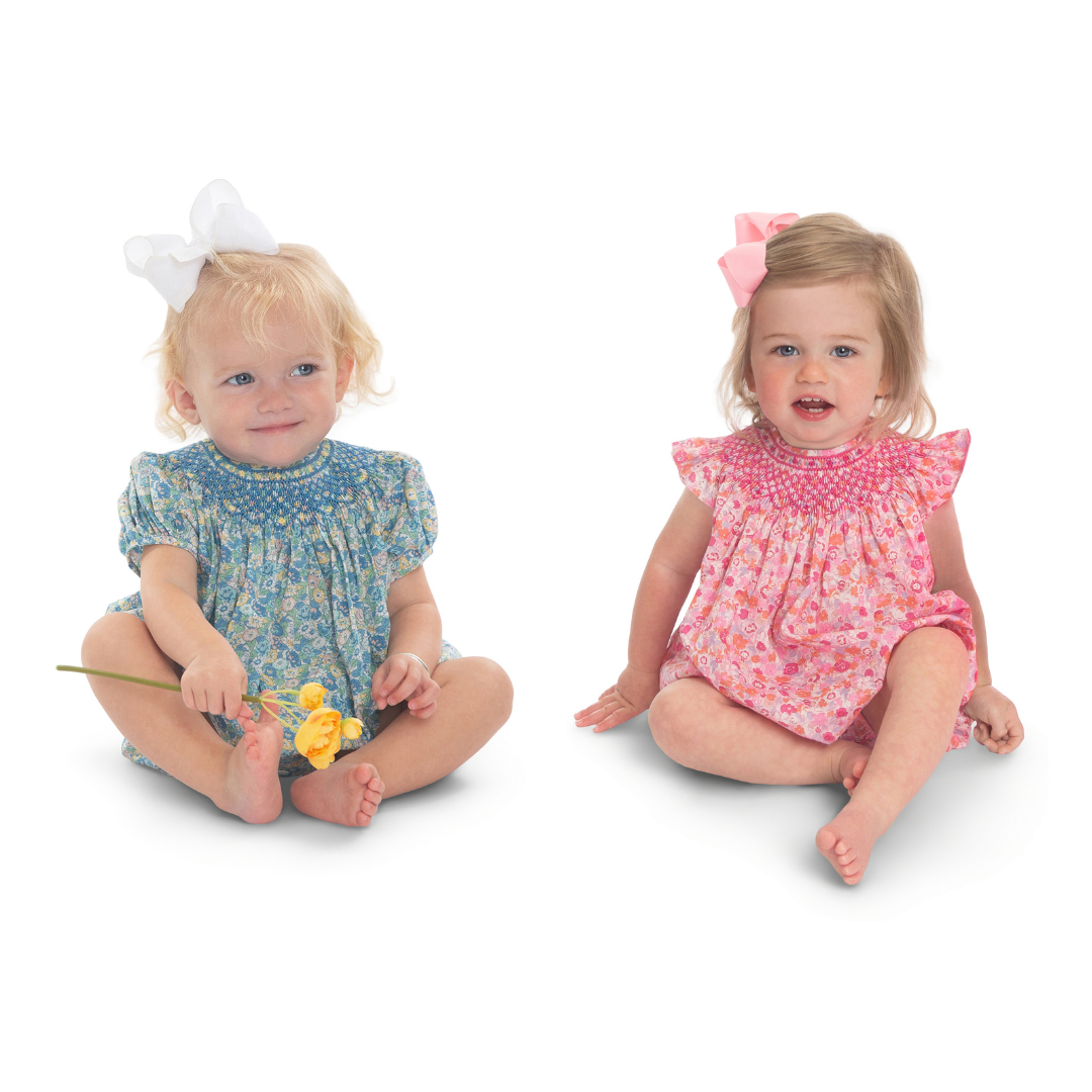 Two baby girls in smocked floral rompers, blue and pink, with hair bows.