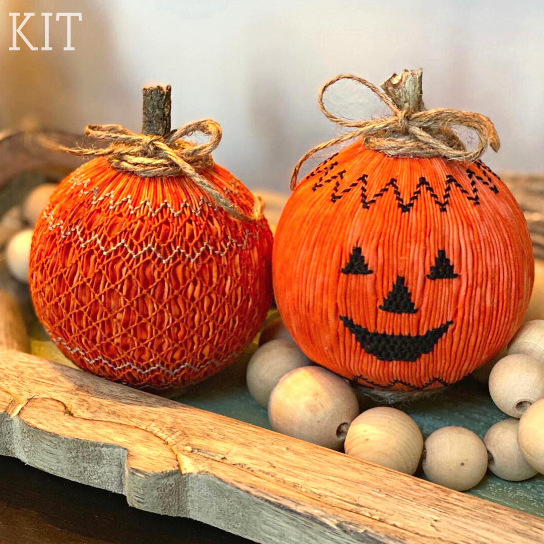 Two smocked orange pumpkins with twine bows, one featuring a cross-stitch jack-o-lantern face.