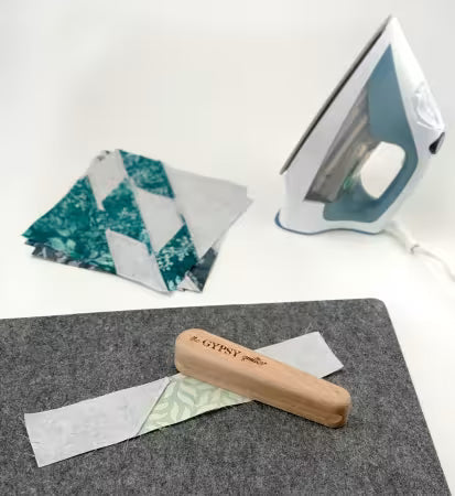 Small wooden clapper tool on gray wool pressing mat with iron and quilt fabric pieces