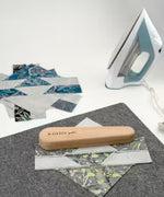 Large wooden clapper tool on felt mat with quilting fabric pieces and iron nearby