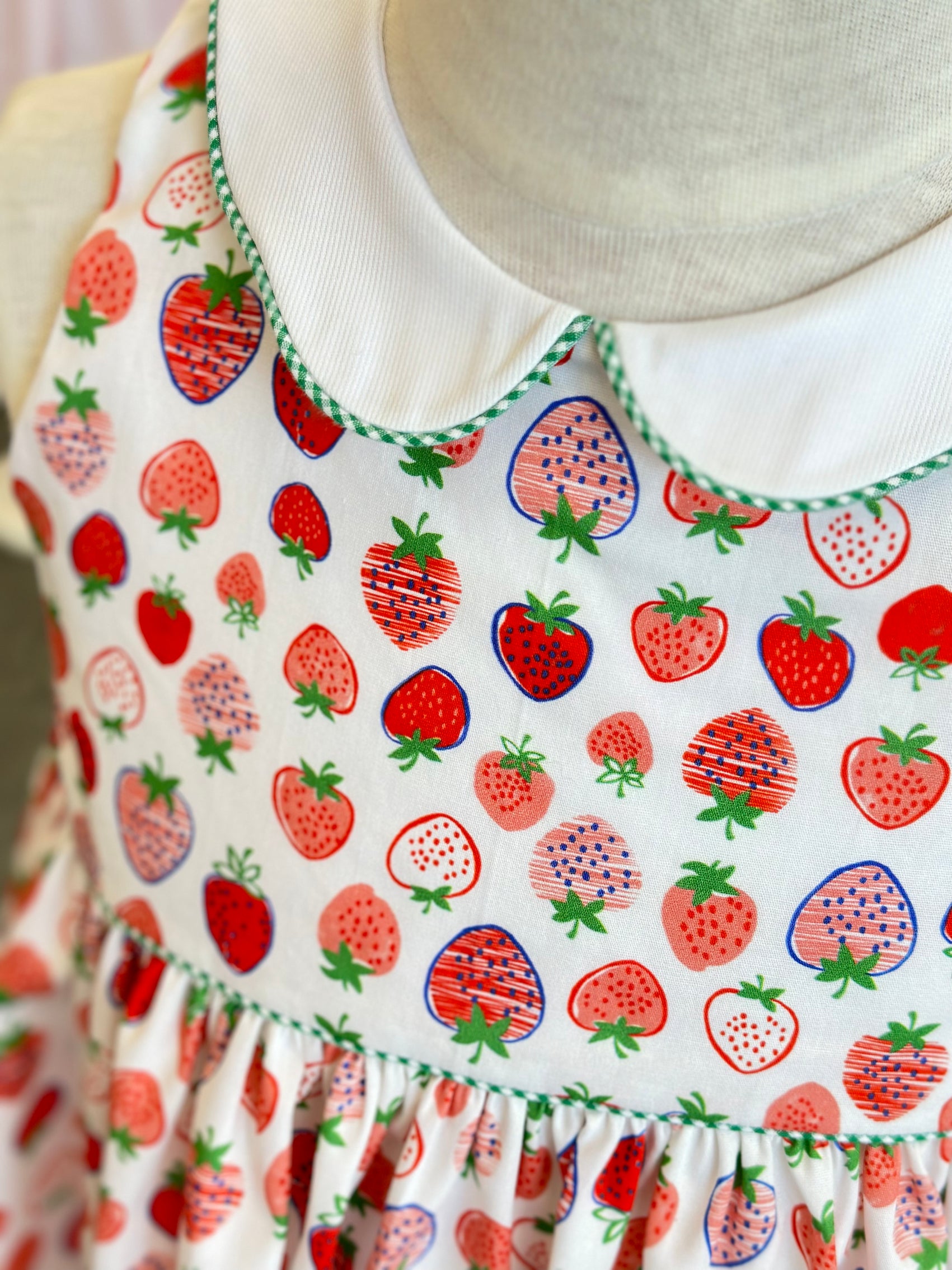 White cotton fabric with red and pink strawberry print, shown as a child's dress with green trim.