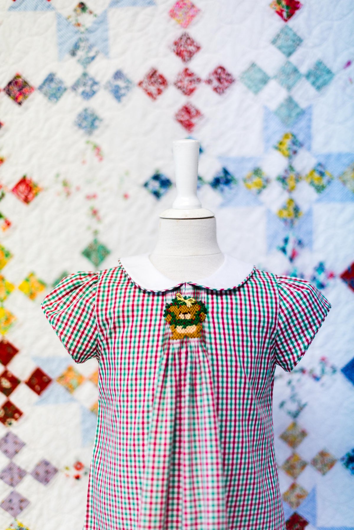 Red and green gingham check fabric displayed on child dress mannequin with quilt backdrop