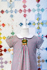 Red and green gingham check fabric displayed on child dress mannequin with quilt backdrop